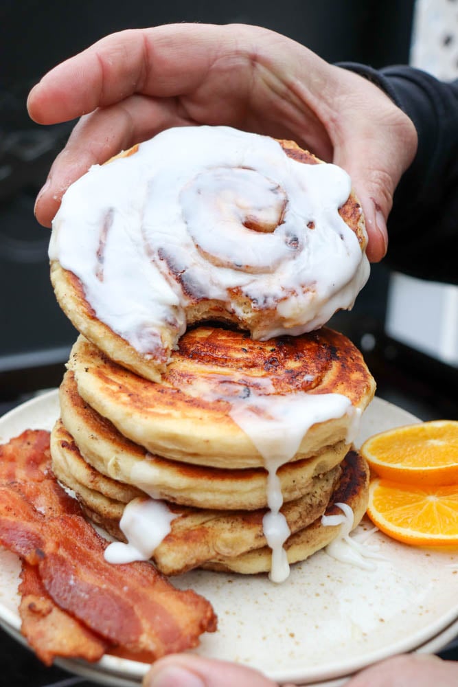 A hand holds a cinnamon roll pancake with icing, stacked on top of more pancakes. On the plate are crispy bacon strips and orange slices. Icing drips down the pancakes.