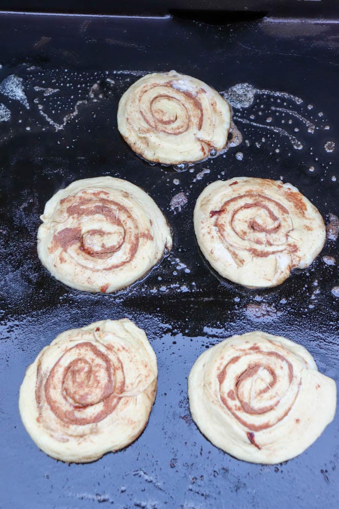 Five cinnamon rolls cooking on a black griddle, with melted butter bubbling around them. The rolls are light golden and swirled with cinnamon filling.