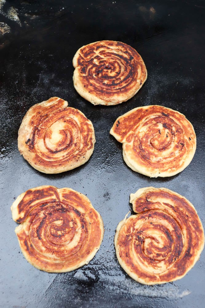 Five round, spiral-shaped flatbreads with golden-brown, crispy surfaces are cooking on a dark, flat griddle. The bread pieces have uneven edges and are arranged in a loose cluster.