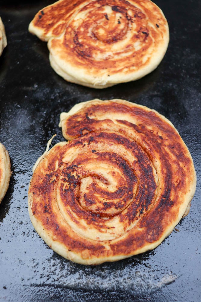 Two spiral-shaped, golden-brown flatbreads are being cooked on a black griddle. The surface of the bread is crispy with visible swirls, and the texture looks flaky and slightly charred in some spots.