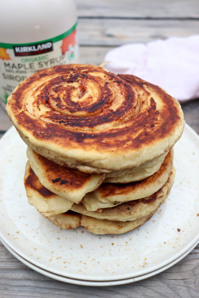 A stack of cinnamon swirl pancakes sits on a speckled plate. In the background, there is a bottle of Kirkland maple syrup and a white cloth on a rustic wooden surface.