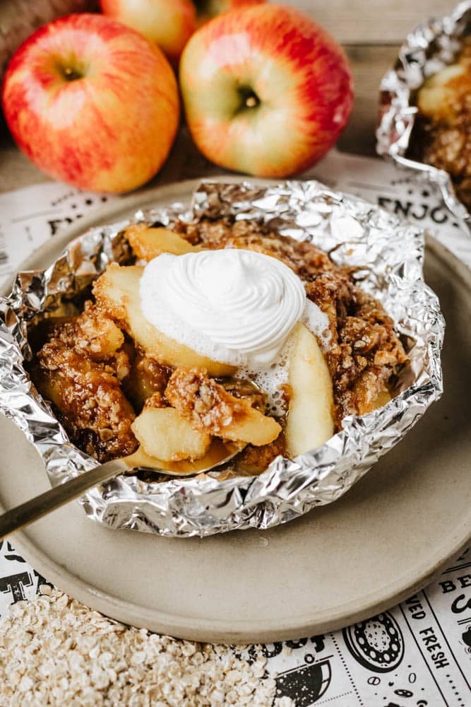 A serving of apple crisp topped with whipped cream sits in foil on a plate. Fresh apples and oats are in the background, suggesting the dessert’s ingredients.