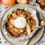 A foil packet filled with baked apple slices and crumble topping, garnished with a dollop of whipped cream, sits on a plate next to a spoon. Fresh apples and oats are in the background.
