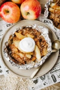 A foil packet filled with baked apple slices and crumble topping, garnished with a dollop of whipped cream, sits on a plate next to a spoon. Fresh apples and oats are in the background.