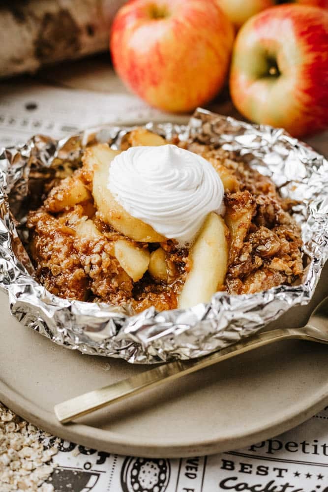 A serving of apple crisp with sliced apples and crumbly topping, baked in foil, topped with a dollop of whipped cream. Fresh apples are in the background, and a fork rests on the plate beside the dessert.