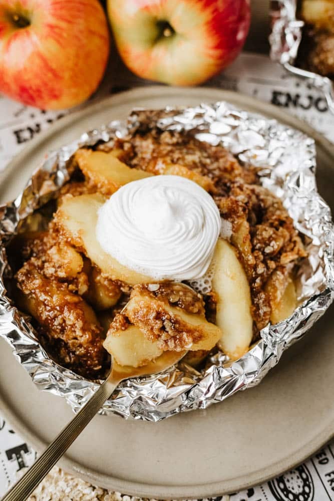 A close-up of baked apple crisp served in a foil packet, topped with a swirl of whipped cream, with fresh apples in the background on a neutral plate.