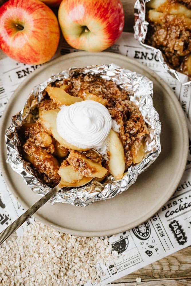 A serving of apple crisp with crumb topping and whipped cream, presented in foil on a plate, with fresh apples and oats scattered nearby.