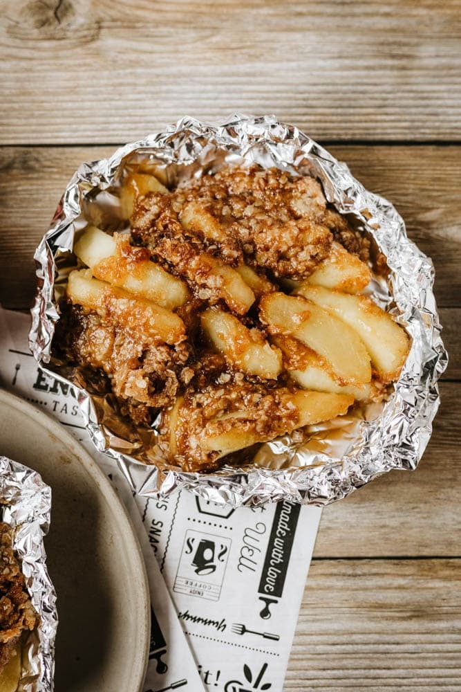 A foil dish filled with a baked dessert made of sliced apples and a crumbly, golden-brown topping, resembling apple crisp, sits on a wooden table.