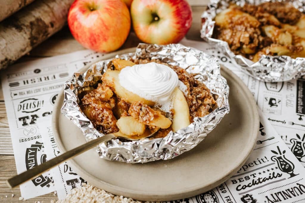 A serving of apple crisp with whipped cream sits in foil on a plate, with fresh apples in the background and a fork resting on the plate.