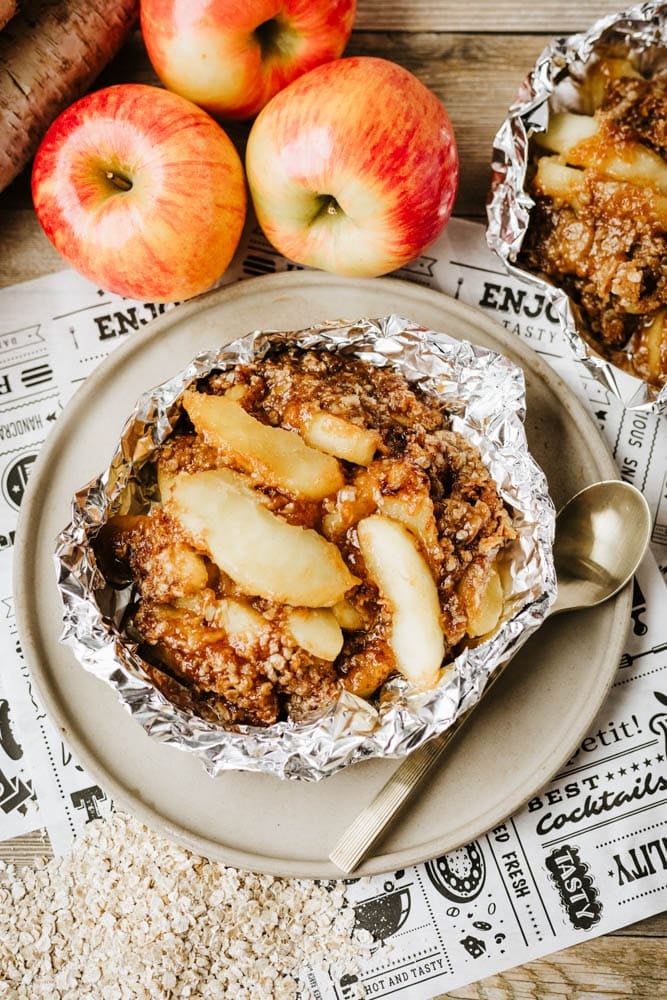 A serving of apple crisp with oat topping in aluminum foil sits on a plate with a spoon. Fresh apples and more foil-wrapped desserts are in the background on a rustic table with decorative paper.
