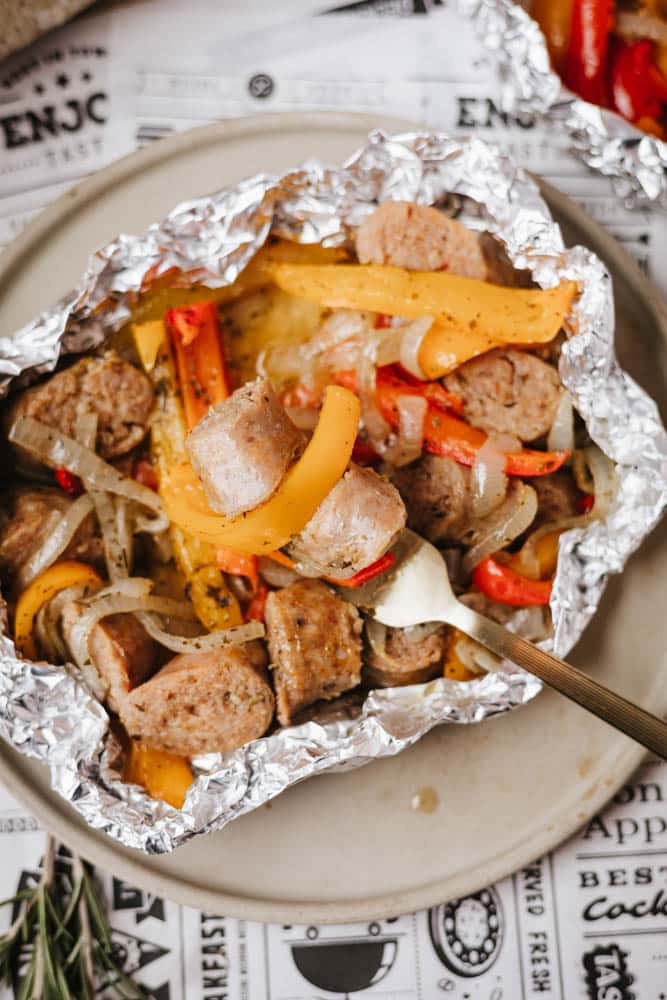 A close-up of sliced sausage, red and yellow bell peppers, and onions cooked in foil, served on a beige plate with a fork. The meal is set on newspaper-style parchment paper.
