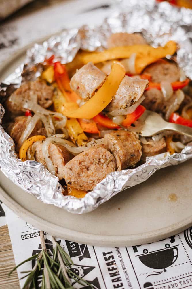 Sliced sausage, yellow and red bell peppers, and onions cooked together in a foil packet, served on a beige plate.