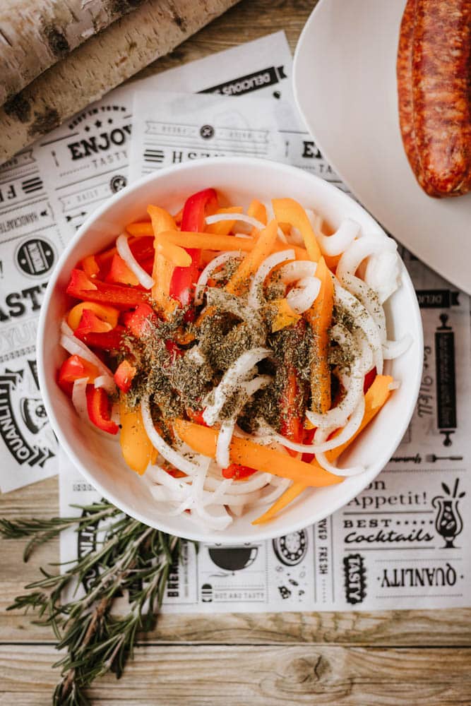 A white bowl filled with sliced red, yellow, and orange bell peppers and white onions, topped with herbs and spices, sits on a decorative paper atop a wooden surface. A sprig of rosemary and a sausage are nearby.