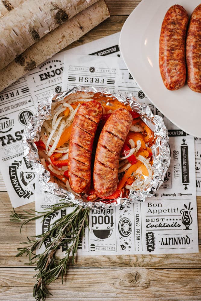 Grilled sausages served on a sheet of foil with sliced onions and bell peppers, garnished with rosemary. Two more sausages are on a white plate nearby, on a wooden table with printed paper underneath.