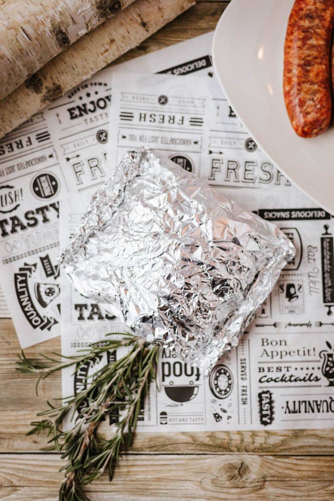 A foil-wrapped food parcel sits on decorative paper with food-related text, next to fresh rosemary sprigs. Part of a white plate with sausages and a birch log are visible on a wooden table.