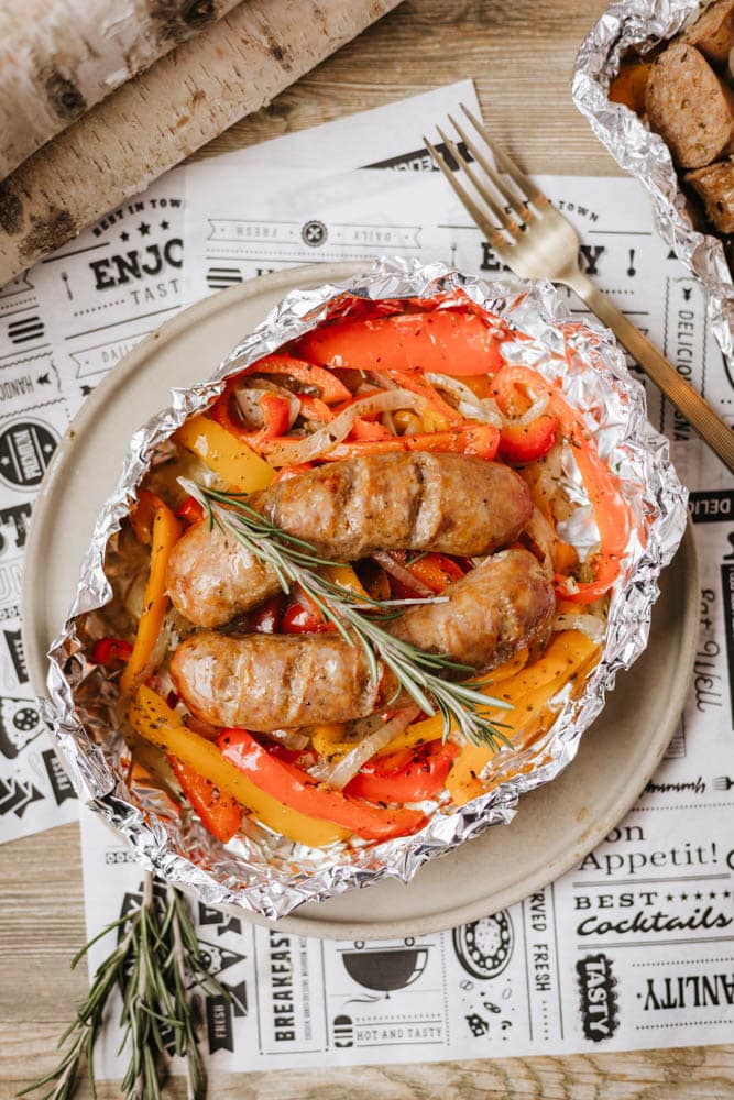 Grilled sausages with sliced red and yellow bell peppers, topped with a sprig of rosemary, served in a foil packet on a plate, with a fork nearby on a rustic table.
