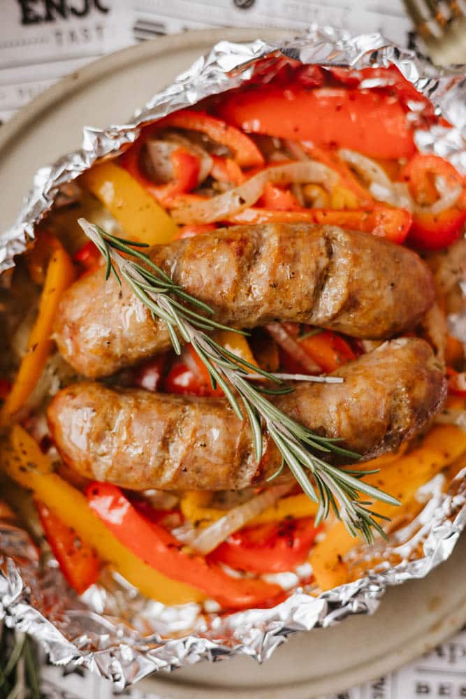Two grilled sausages served on a bed of sautéed red and yellow bell peppers and onions, garnished with a sprig of rosemary, all placed in a foil packet on a plate.