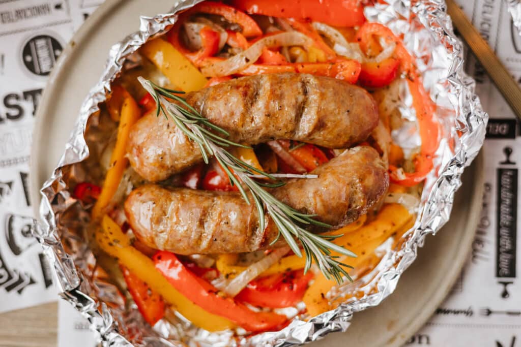 Three grilled sausages topped with a sprig of rosemary sit on a bed of sliced red and yellow bell peppers and onions, all served in a foil packet on a round plate.