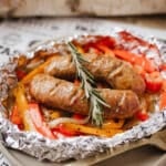 Grilled sausages served on a bed of sliced red and yellow bell peppers and onions, all placed in a foil packet and garnished with a sprig of rosemary.