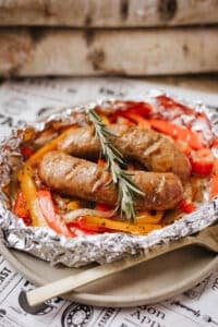 Grilled sausages served on a bed of sliced red and yellow bell peppers and onions, all placed in a foil packet and garnished with a sprig of rosemary.