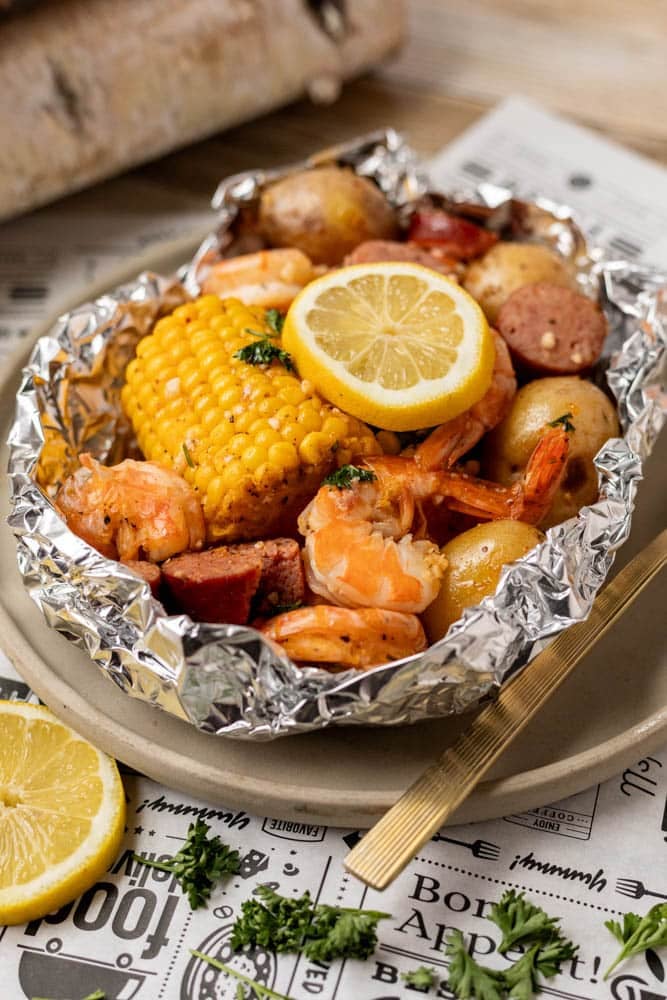 A foil packet filled with shrimp, corn on the cob, baby potatoes, sausage slices, and garnished with fresh parsley and lemon slices, served on a plate.