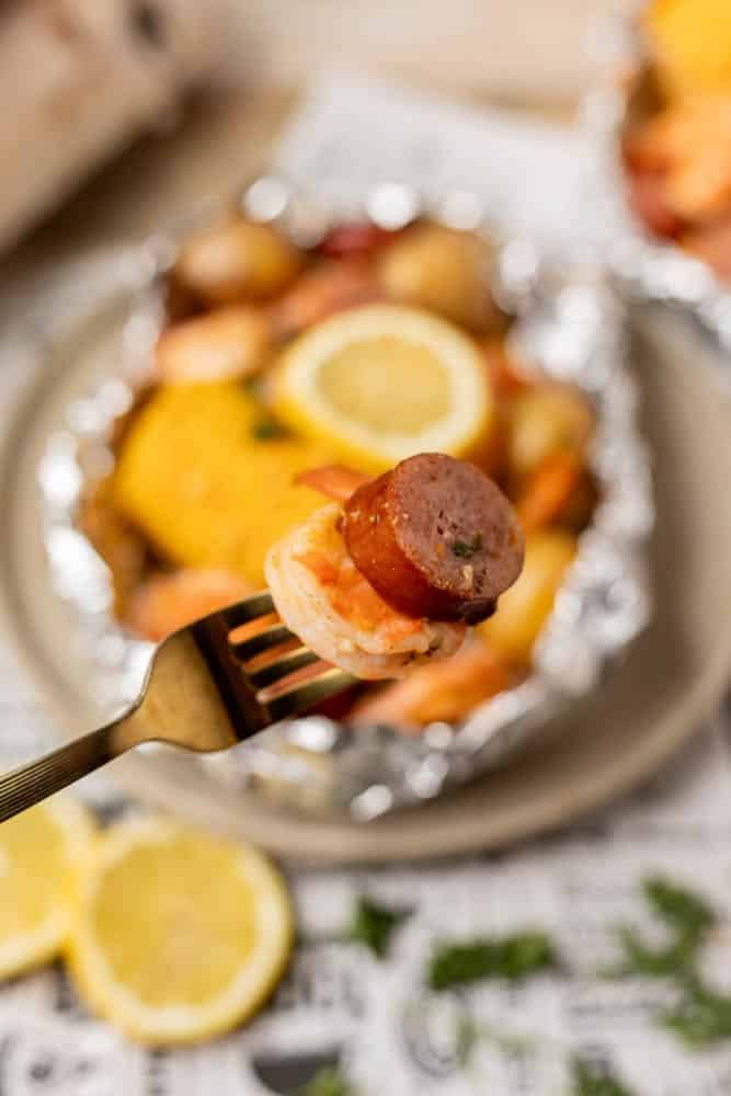 A close-up of a fork holding a slice of sausage and shrimp, with a foil packet meal containing corn, potatoes, shrimp, and a lemon slice blurred in the background.