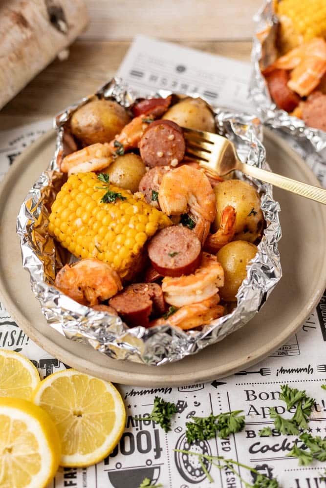 Foil packet containing shrimp, sausage slices, baby potatoes, and a corn cob, garnished with herbs on a plate. A gold fork is in the packet. Lemon slices and parsley are on the side, and a food-themed paper is underneath.