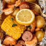 A foil packet filled with shrimp, sliced sausage, corn on the cob, baby potatoes, and herbs, topped with a lemon slice and served with a gold fork.