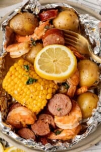 A foil packet filled with shrimp, sliced sausage, corn on the cob, baby potatoes, and herbs, topped with a lemon slice and served with a gold fork.
