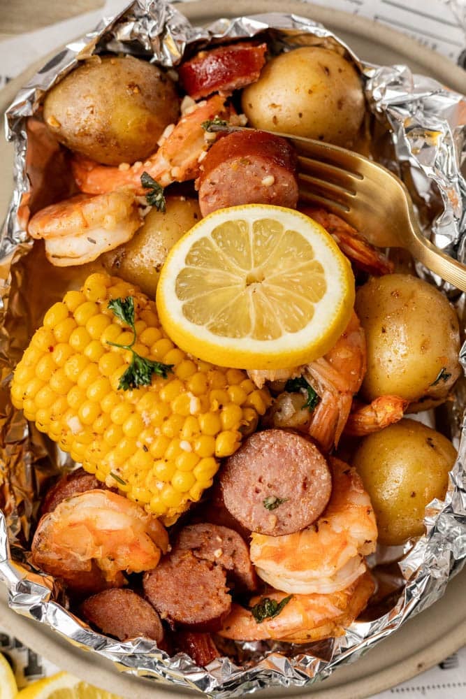 A foil packet filled with shrimp, sliced sausage, corn on the cob, baby potatoes, and herbs, topped with a lemon slice and served with a gold fork.