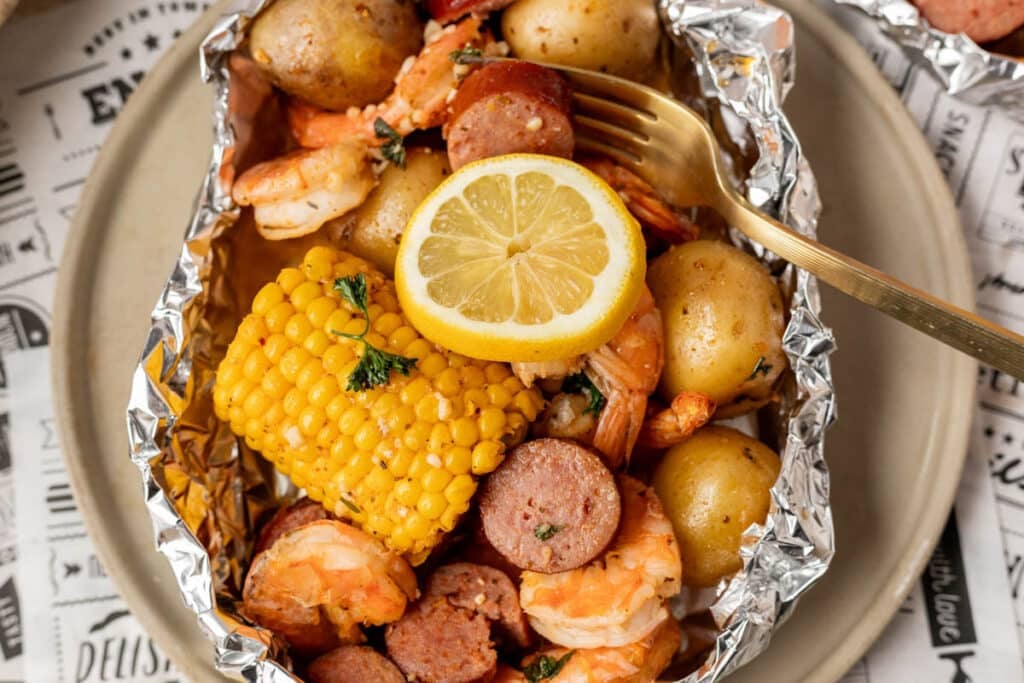 A foil packet filled with shrimp, sliced sausage, baby potatoes, corn on the cob, and garnished with lemon slices and parsley, served on a beige plate with a gold fork.
