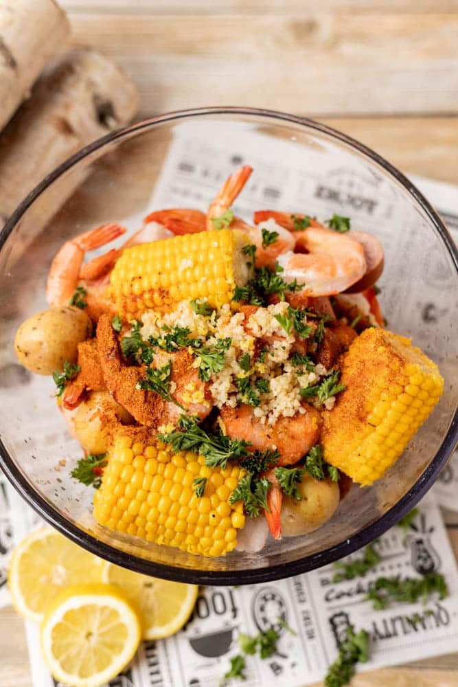 A glass bowl filled with shrimp, corn on the cob, baby potatoes, and garnished with chopped parsley and seasoning. Lemon slices are placed beside the bowl on a newspaper-style surface.