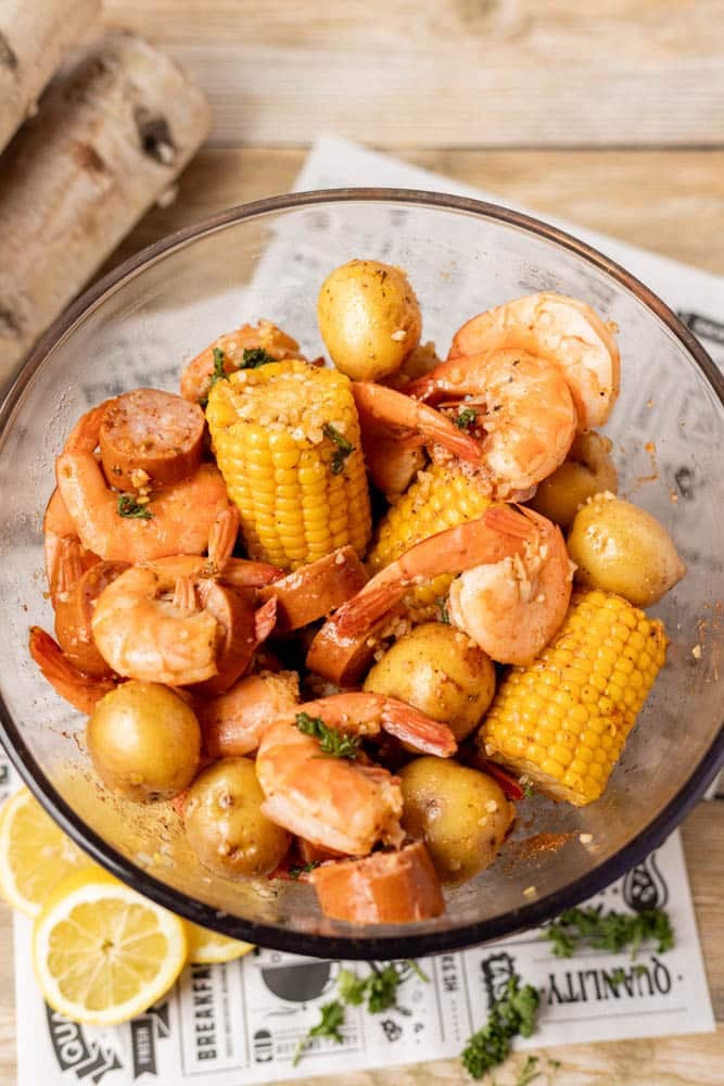 A glass bowl filled with shrimp, corn on the cob, baby potatoes, and sliced sausage, garnished with herbs. A few lemon slices and some wooden planks are nearby on the table.