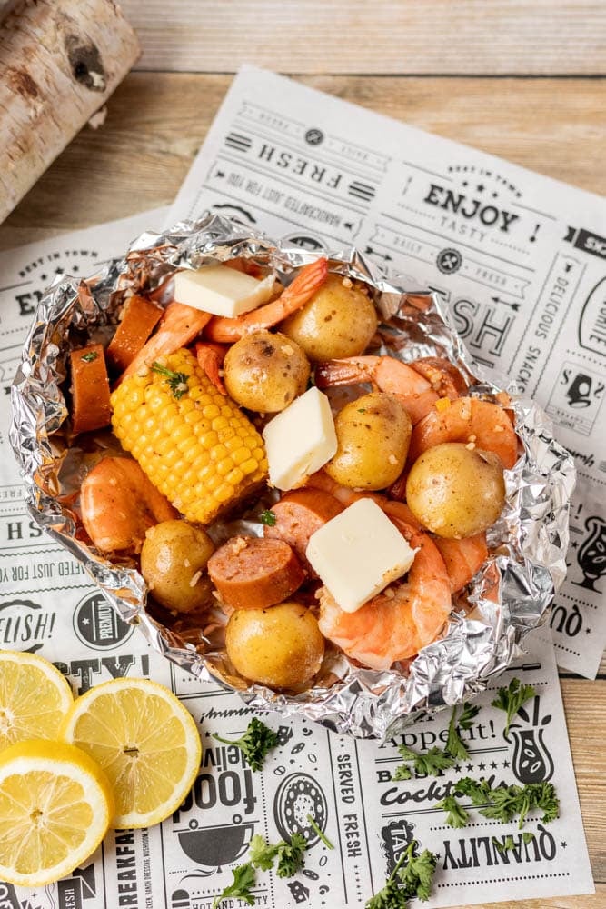 A foil packet filled with shrimp, small potatoes, corn on the cob, sausage slices, and pats of butter sits on newspaper-style parchment, with lemon slices and parsley garnish on the side.