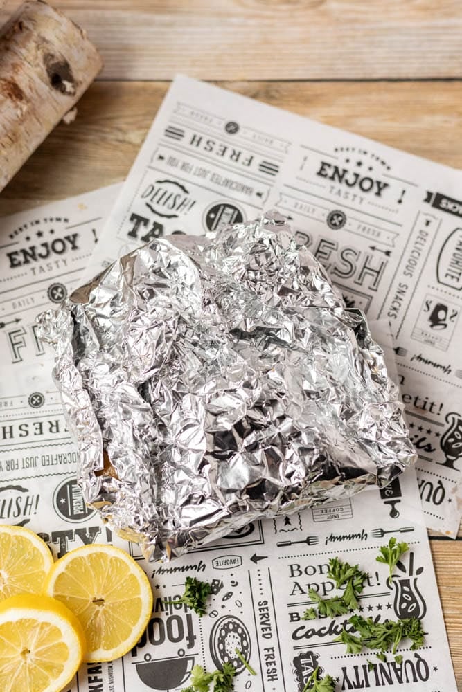 A crumpled foil-wrapped food packet sits on top of decorative paper with food-related words, surrounded by fresh lemon slices and sprigs of herbs on a wooden surface.