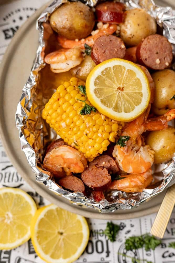 A foil packet filled with shrimp, sliced sausage, small potatoes, and a corn cob, garnished with fresh parsley and two lemon slices; more lemon slices are on the plate beside the packet.