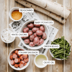 Top-down view of labeled ingredients on a wooden table: a bowl of baby potatoes, cut green beans, sliced white onions, Italian sausage pieces, a small bowl of seasoning, and a cup of olive oil.