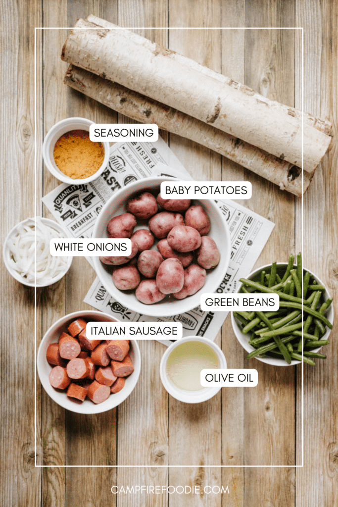 Top-down view of labeled ingredients on a wooden table: a bowl of baby potatoes, cut green beans, sliced white onions, Italian sausage pieces, a small bowl of seasoning, and a cup of olive oil.