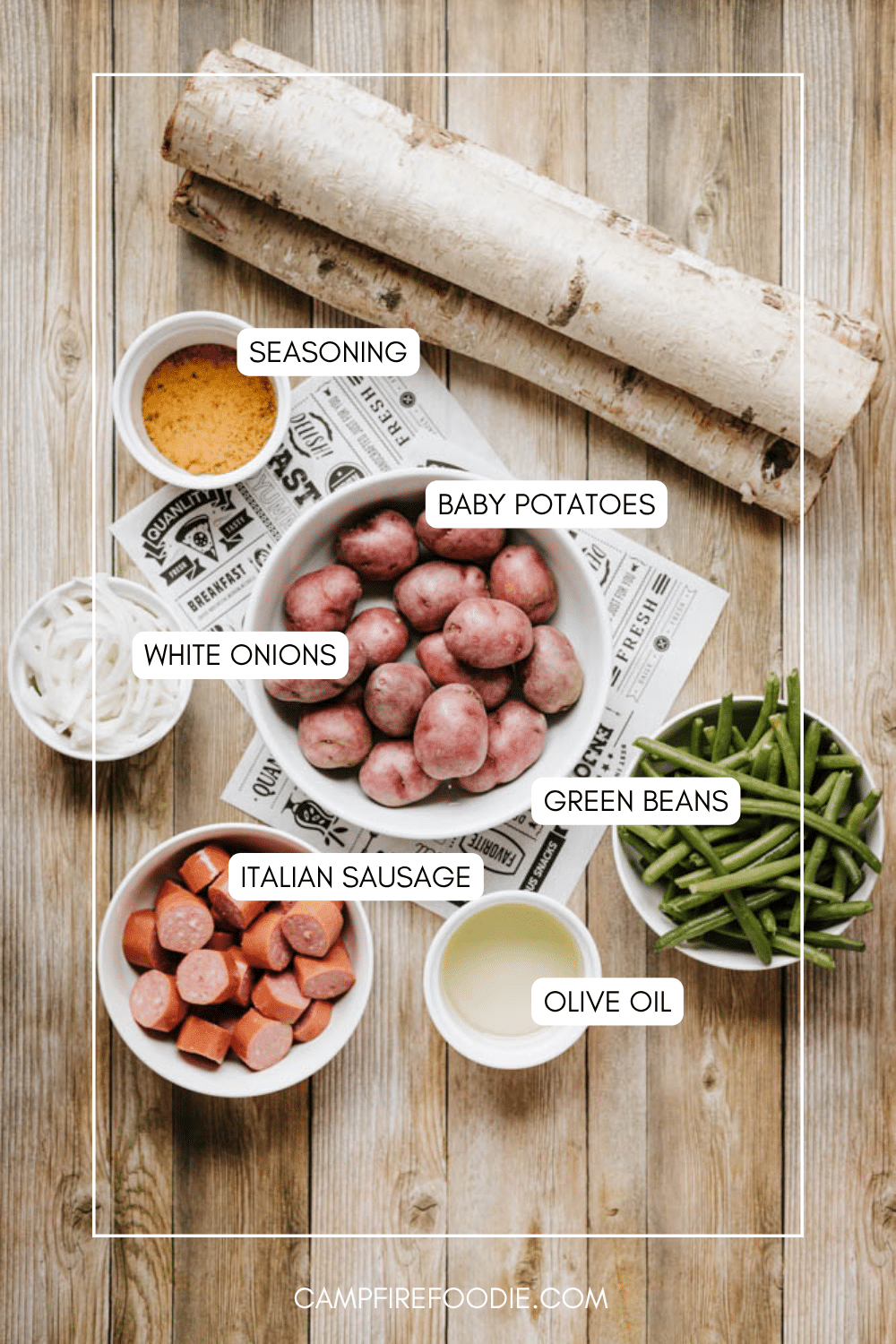 Top-down view of labeled ingredients on a wooden table: a bowl of baby potatoes, cut green beans, sliced white onions, Italian sausage pieces, a small bowl of seasoning, and a cup of olive oil.