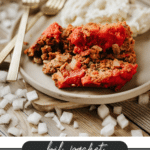 A plate with two slices of meatloaf topped with tomato sauce, served with mashed potatoes. Gold utensils lie beside the plate; diced onions are scattered on the wooden table. Text reads: Foil Packet Meatloaf CampfireFoodie.com.