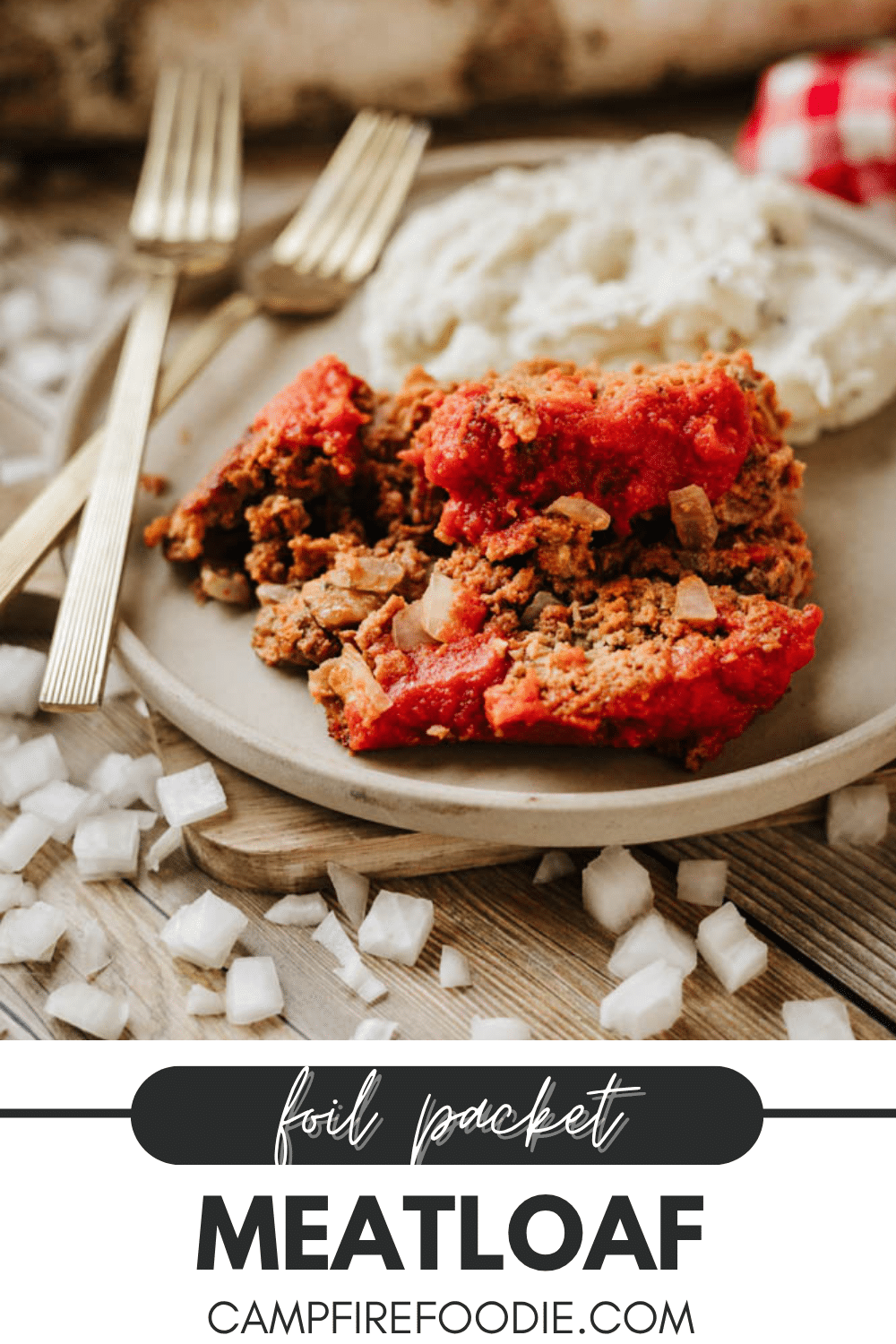 A plate with two slices of meatloaf topped with tomato sauce, served with mashed potatoes. Gold utensils lie beside the plate; diced onions are scattered on the wooden table. Text reads: Foil Packet Meatloaf CampfireFoodie.com.