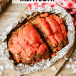 A loaf of meatloaf topped with red sauce sits in an open foil packet on a wooden table, surrounded by chopped onions and a red checkered cloth. Text reads: foil packet MEATLOAF and campfirefoodie.com.