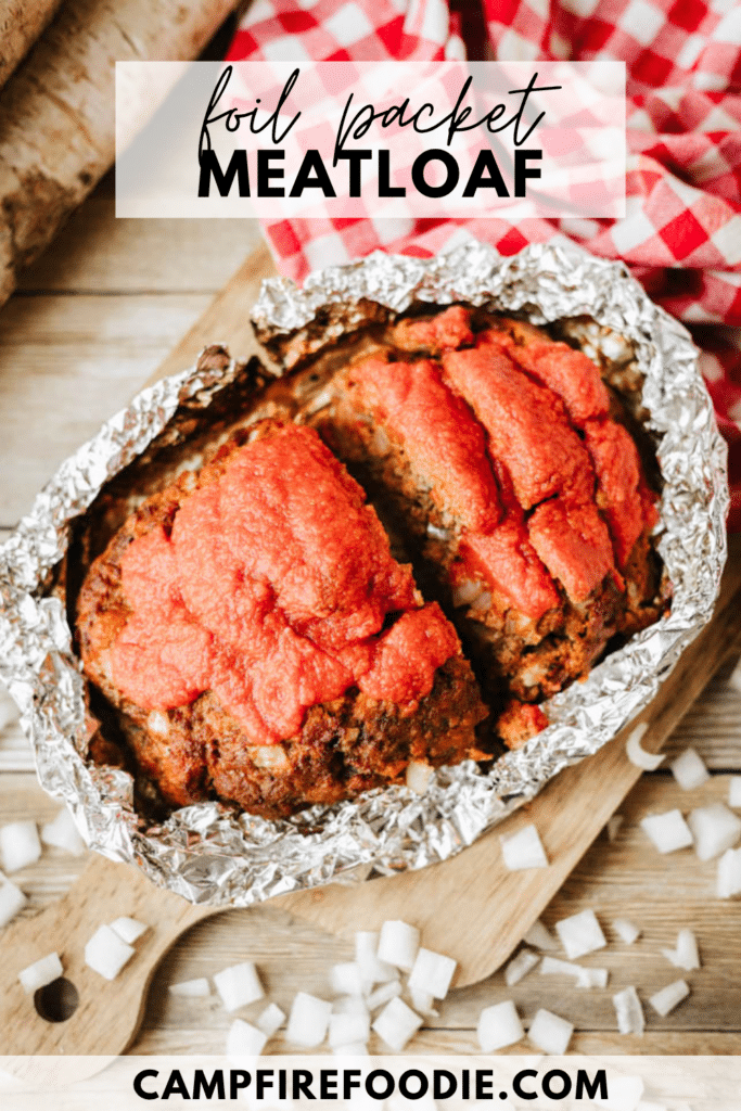 A loaf of meatloaf topped with red sauce sits in an open foil packet on a wooden table, surrounded by chopped onions and a red checkered cloth. Text reads: foil packet MEATLOAF and campfirefoodie.com.