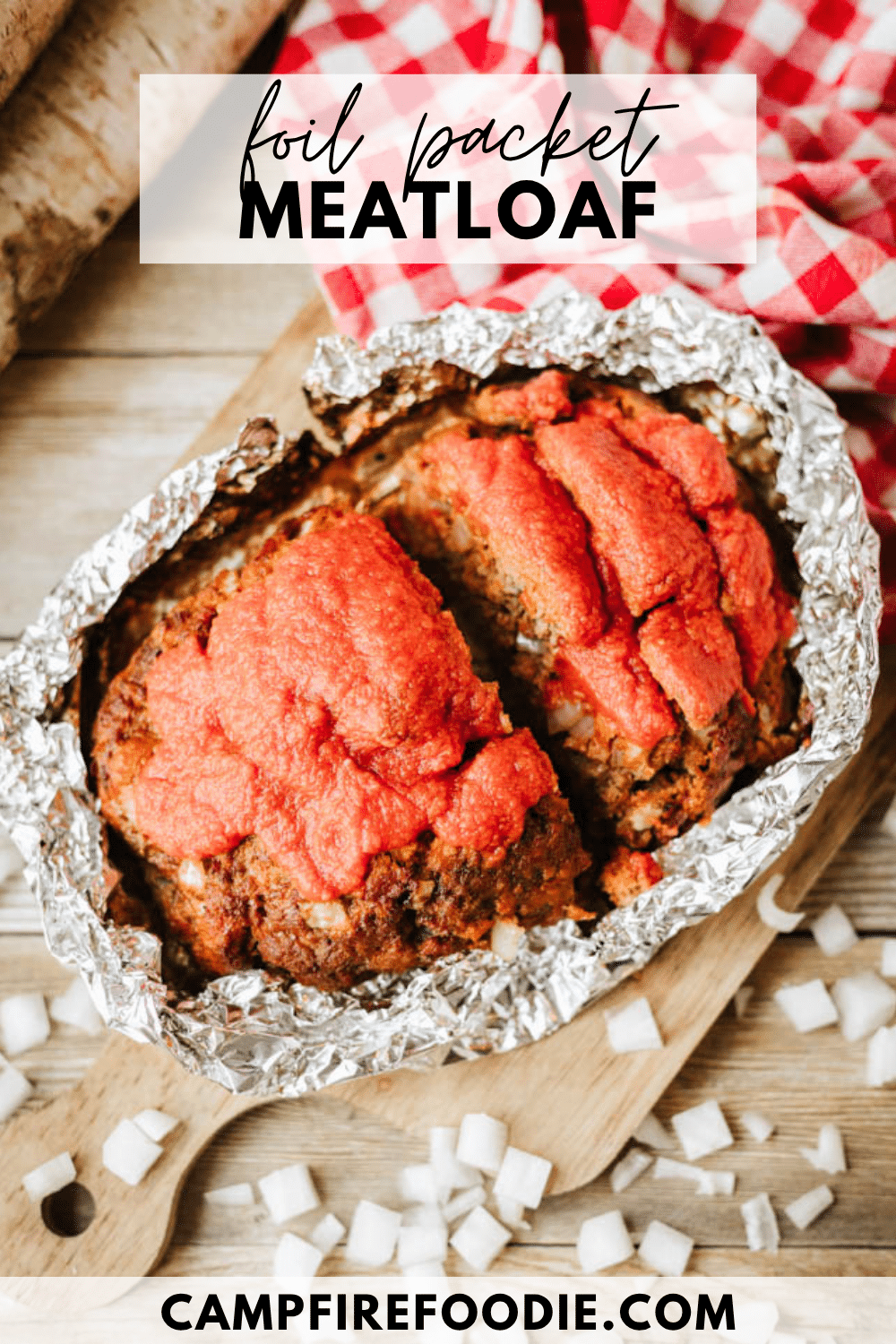 A loaf of meatloaf topped with red sauce sits in an open foil packet on a wooden table, surrounded by chopped onions and a red checkered cloth. Text reads: foil packet MEATLOAF and campfirefoodie.com.