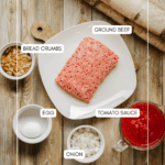 Overhead view of a plate with raw ground beef, surrounded by bowls containing bread crumbs, a whole egg, chopped onion, and tomato sauce on a wooden surface. Each ingredient is labeled.