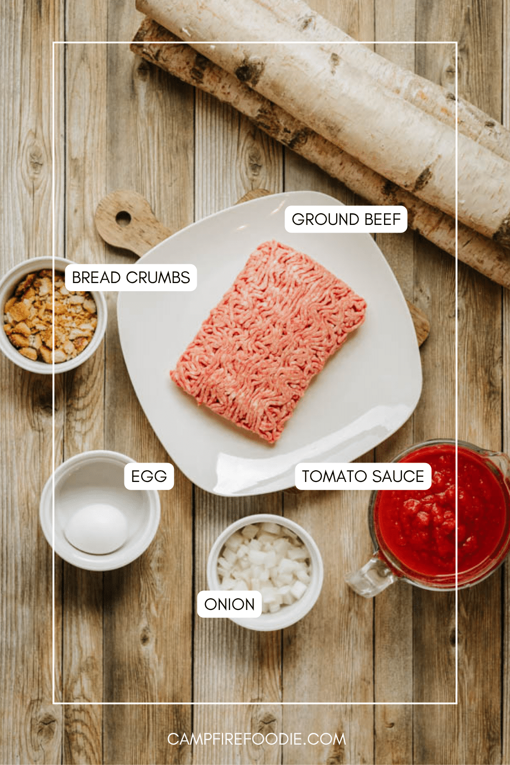 Overhead view of a plate with raw ground beef, surrounded by bowls containing bread crumbs, a whole egg, chopped onion, and tomato sauce on a wooden surface. Each ingredient is labeled.