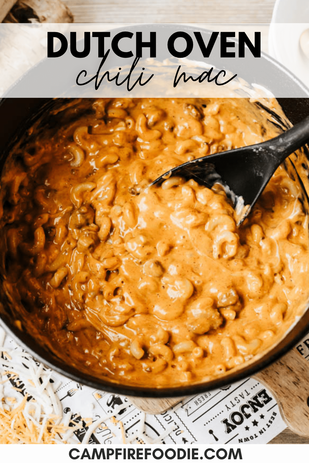 A black Dutch oven filled with creamy chili mac and a black spoon, with Dutch Oven Chili Mac text above. The image is branded with campfirefoodie.com at the bottom.
