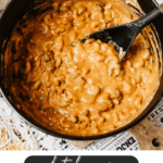 A black Dutch oven filled with creamy chili mac topped with melted cheese sits on a table. A black spoon is stirring the pasta. Text reads: Dutch Oven Chili Mac, campfirefoodie.com.