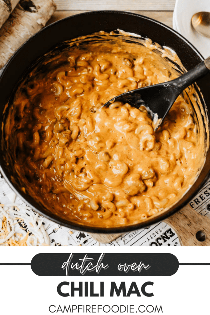 A black Dutch oven filled with creamy chili mac topped with melted cheese sits on a table. A black spoon is stirring the pasta. Text reads: Dutch Oven Chili Mac, campfirefoodie.com.