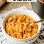 A white bowl filled with creamy chili mac sits on a patterned surface with a spoon inside. Behind it is a black Dutch oven. Text on the image reads dutch oven chili mac and campfirefoodie.com.